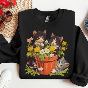 May include: Black sweatshirt with a graphic of three kittens in a flower pot. The kittens are black and white with green, yellow, and white flowers around them.