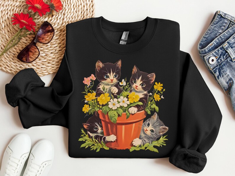 May include: Black sweatshirt with a graphic of three kittens in a flower pot. The kittens are black and white with green, yellow, and white flowers around them.