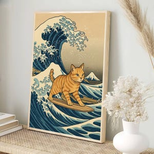 May include: A framed art print featuring a tabby cat surfing a wave, inspired by Japanese art. The cat is orange with black stripes, riding a wooden surfboard on a blue and white wave. The background includes a mountain and a light beige sky.