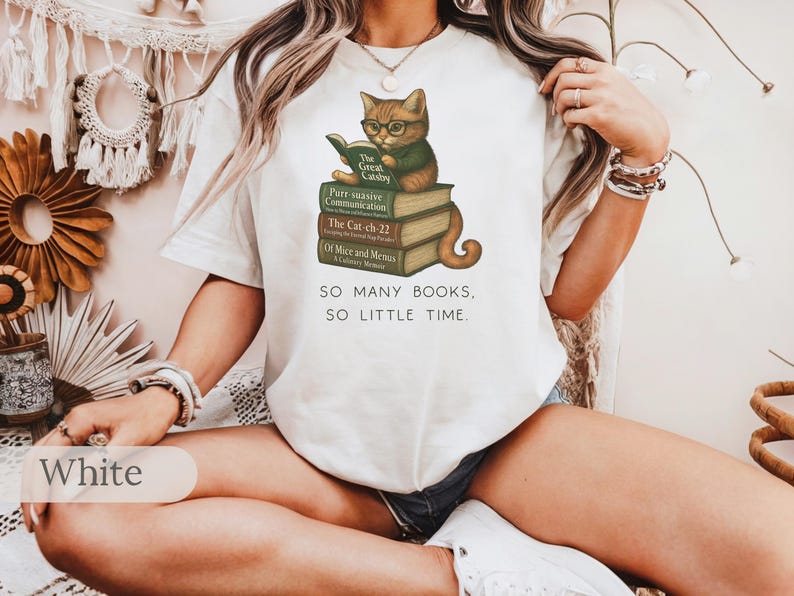 K&ouml;nnte beinhalten: Wei&szlig;es T-Shirt mit einem Grafikdruck eines Katzen mit Brille, das auf einem Stapel B&uuml;cher sitzt. Der Text auf dem Shirt lautet "SO MANY BOOKS, SO LITTLE TIME."