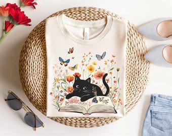 May include: A white t-shirt with a black cat reading a book surrounded by colorful flowers and butterflies. The book has the words "Never Stop" on the cover.
