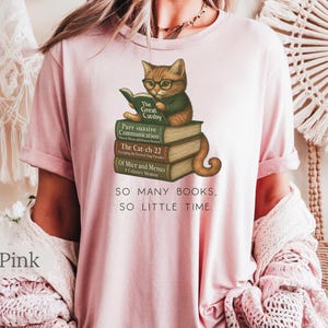 K&ouml;nnte beinhalten: Ein T-Shirt in zartem Rosa mit einem Grafikdruck eines Katers, der ein Buch auf einem Stapel B&uuml;cher liest. Der Text auf dem T-Shirt lautet "SO MANY BOOKS, SO LITTLE TIME."