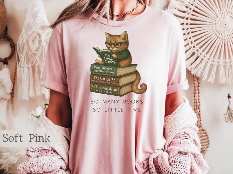 K&ouml;nnte beinhalten: Ein T-Shirt in zartem Rosa mit einem Grafikdruck eines Katers, der ein Buch auf einem Stapel B&uuml;cher liest. Der Text auf dem T-Shirt lautet "SO MANY BOOKS, SO LITTLE TIME."
