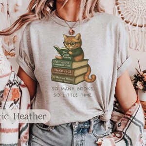 K&ouml;nnte beinhalten: Ein heidefarbenes T-Shirt mit einem Aufdruck eines Katers, der einen Stapel B&uuml;cher liest. Der Text auf dem Shirt lautet "SO MANY BOOKS, SO LITTLE TIME."