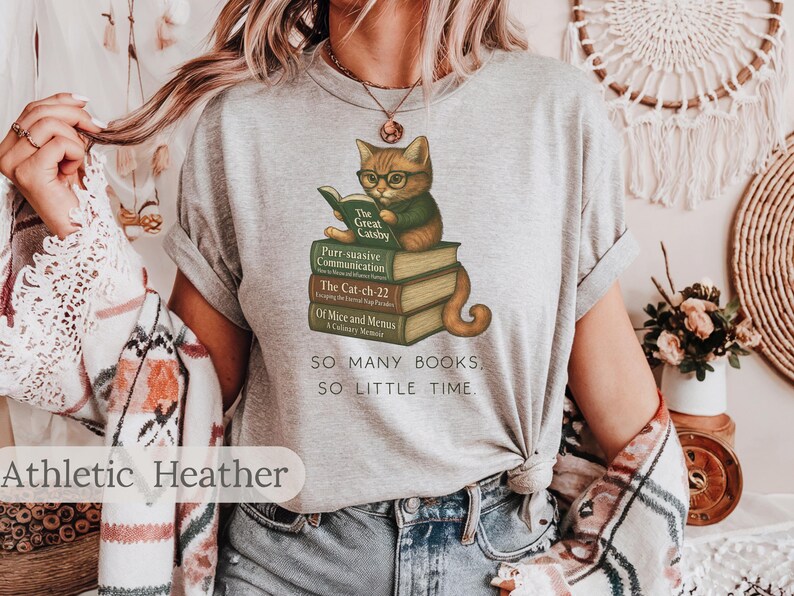 K&ouml;nnte beinhalten: Ein heidefarbenes T-Shirt mit einem Aufdruck eines Katers, der einen Stapel B&uuml;cher liest. Der Text auf dem Shirt lautet "SO MANY BOOKS, SO LITTLE TIME."