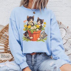 May include: A light blue sweatshirt with a graphic of three kittens in a flower pot. The kittens are black and white with yellow flowers around them.
