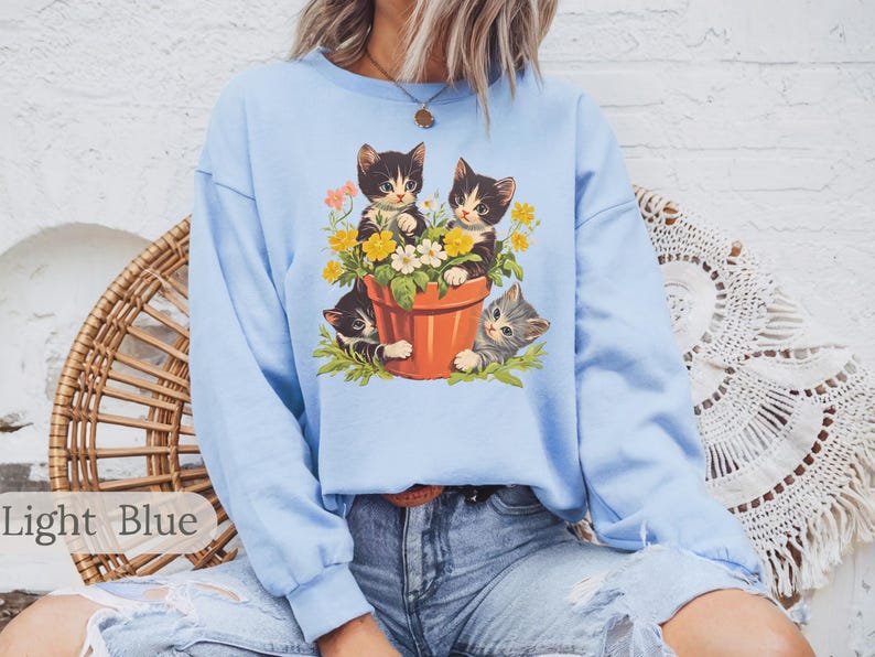 May include: A light blue sweatshirt with a graphic of three kittens in a flower pot. The kittens are black and white with yellow flowers around them.