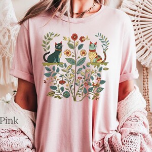 May include: A soft pink t-shirt with a floral design featuring two cats. The cats are stylized and have a folk art aesthetic. The design includes flowers, leaves, and vines.