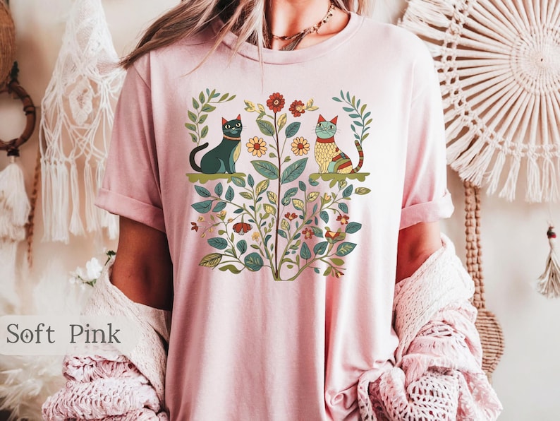 May include: A soft pink t-shirt with a floral design featuring two cats. The cats are stylized and have a folk art aesthetic. The design includes flowers, leaves, and vines.