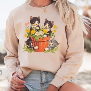 May include: A beige sweatshirt with a vintage illustration of three kittens in a flower pot. The kittens are black and white with green, yellow, and white flowers around them.