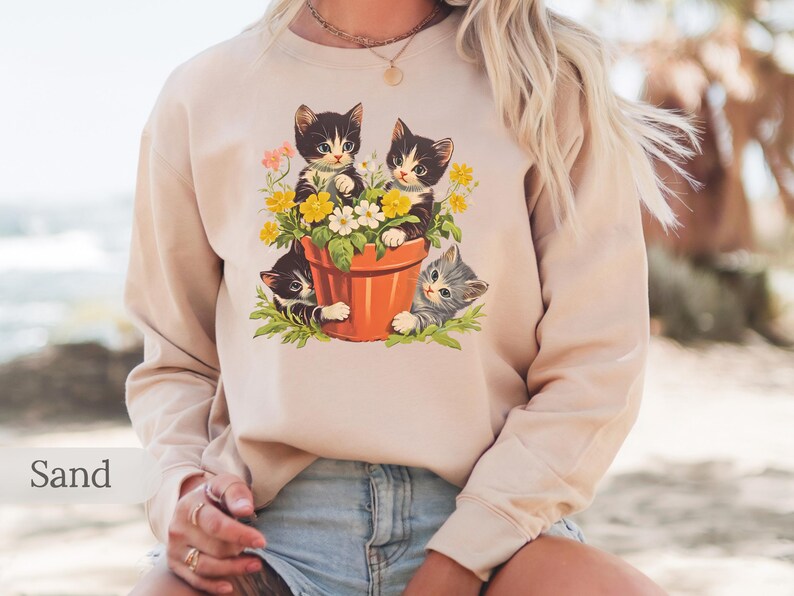May include: A beige sweatshirt with a vintage illustration of three kittens in a flower pot. The kittens are black and white with green, yellow, and white flowers around them.
