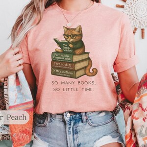 K&ouml;nnte beinhalten: Ein pinkfarbenes T-Shirt mit einem Motiv eines Katzen, das eine Brille tr&auml;gt und ein Buch liest. Die Katze sitzt auf einem Stapel B&uuml;cher mit Titeln wie "The Great Gatsby", "Purr suasive Communication", "The Cat-ch-22" und "Of Mice and Menus". Der Text "SO MANY BOOKS, SO LITTLE TIME." ist unter der Katze und den B&uuml;chern gedruckt.