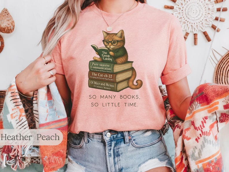 K&ouml;nnte beinhalten: Ein pinkfarbenes T-Shirt mit einem Motiv eines Katzen, das eine Brille tr&auml;gt und ein Buch liest. Die Katze sitzt auf einem Stapel B&uuml;cher mit Titeln wie "The Great Gatsby", "Purr suasive Communication", "The Cat-ch-22" und "Of Mice and Menus". Der Text "SO MANY BOOKS, SO LITTLE TIME." ist unter der Katze und den B&uuml;chern gedruckt.
