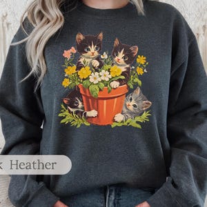 May include: A dark heather grey sweatshirt with a graphic of three black and white kittens peeking out of a flower pot with colorful flowers. The text "Dark Heather" is printed on the bottom left corner of the sweatshirt.