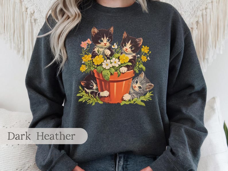 May include: A dark heather grey sweatshirt with a graphic of three black and white kittens peeking out of a flower pot with colorful flowers. The text "Dark Heather" is printed on the bottom left corner of the sweatshirt.