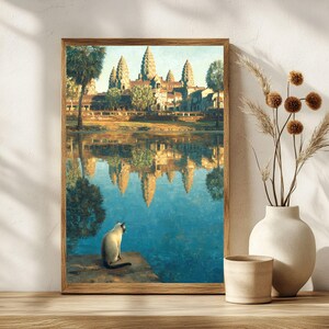 May include: A framed art print showing the Angkor Wat temple complex reflected in water. A Siamese cat sits on a stone ledge, observing the scene. The print is in a wooden frame, with a calm, artistic style. The colour palette is dominated by blues and browns.