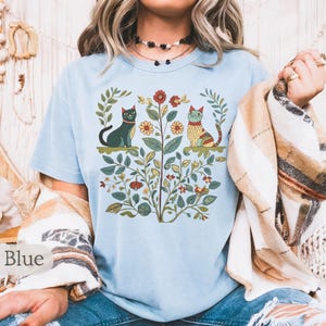 May include: A light blue t-shirt with a floral design featuring two cats. One cat is black and the other is orange and yellow. The cats are surrounded by flowers and leaves.