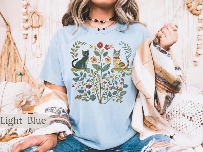 May include: A light blue t-shirt with a floral design featuring two cats. One cat is black and the other is orange and yellow. The cats are surrounded by flowers and leaves.