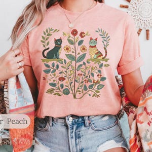 May include: A pink t-shirt with a graphic print of two cats sitting in a garden with flowers and leaves. The text "Heather Peach" is visible at the bottom of the image.