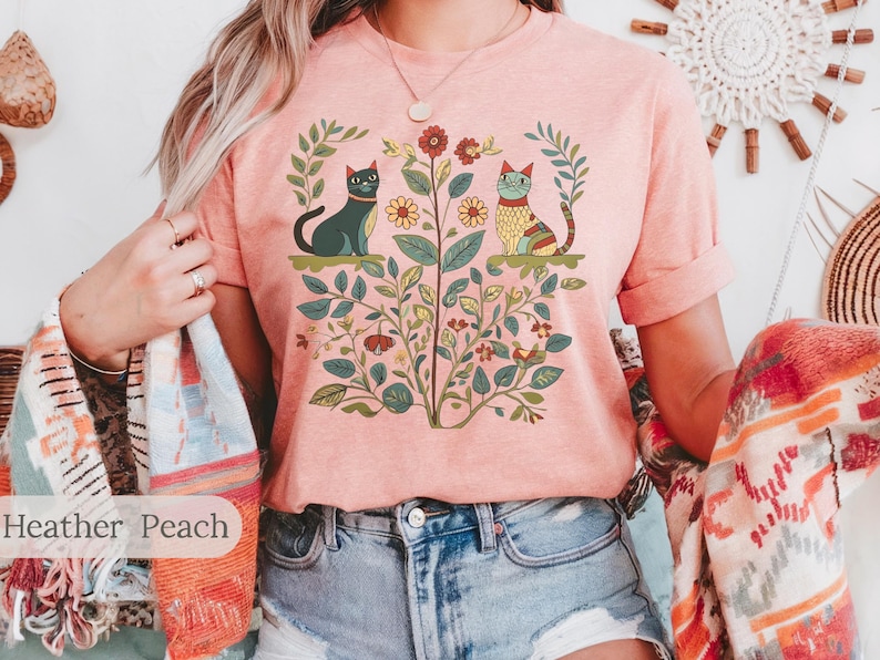 May include: A pink t-shirt with a graphic print of two cats sitting in a garden with flowers and leaves. The text "Heather Peach" is visible at the bottom of the image.