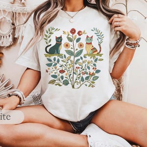 May include: White t-shirt with a floral design featuring two cats. The cats are stylized and have a folk art aesthetic. The shirt has short sleeves and a crew neck.