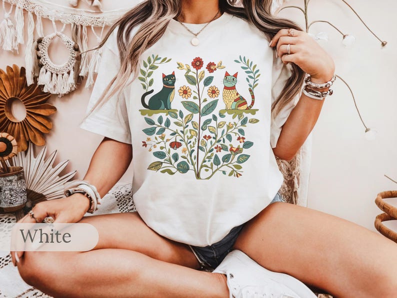 May include: White t-shirt with a floral design featuring two cats. The cats are stylized and have a folk art aesthetic. The shirt has short sleeves and a crew neck.