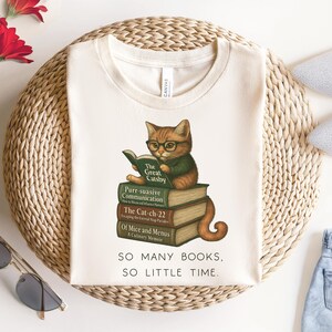 K&ouml;nnte beinhalten: Ein wei&szlig;es T-Shirt mit einem Motiv eines Katzen, das eine Brille tr&auml;gt und ein Buch liest. Die Katze sitzt auf einem Stapel B&uuml;cher mit den Titeln "The Great Gatsby", "Purr-suasive Communication", "The Cat-ch-22" und "Of Mice and Menus: A Culinary Memoir". Der Text "SO MANY BOOKS, SO LITTLE TIME." ist unter dem Motiv gedruckt.