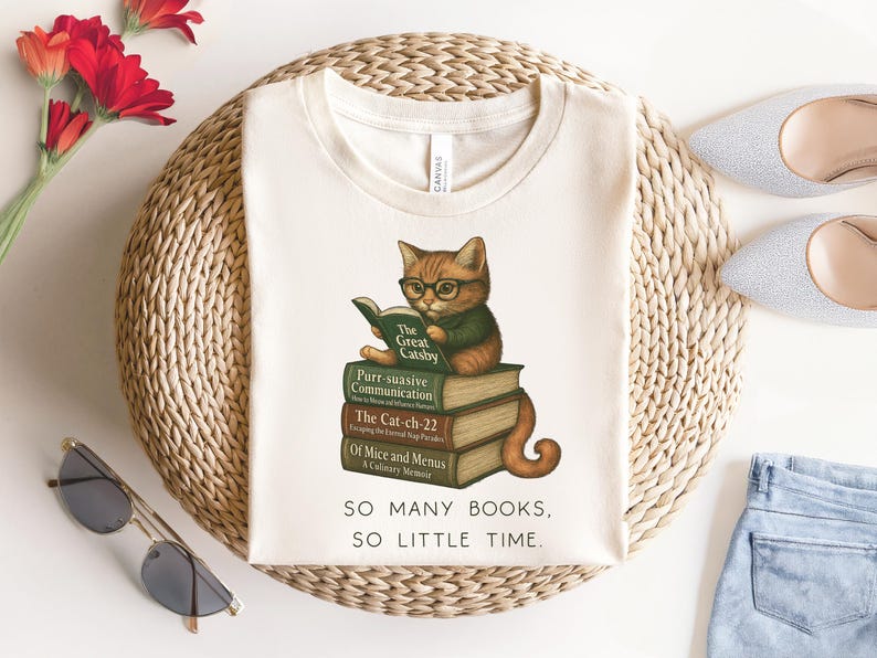 K&ouml;nnte beinhalten: Ein wei&szlig;es T-Shirt mit einem Motiv eines Katzen, das eine Brille tr&auml;gt und ein Buch liest. Die Katze sitzt auf einem Stapel B&uuml;cher mit den Titeln "The Great Gatsby", "Purr-suasive Communication", "The Cat-ch-22" und "Of Mice and Menus: A Culinary Memoir". Der Text "SO MANY BOOKS, SO LITTLE TIME." ist unter dem Motiv gedruckt.