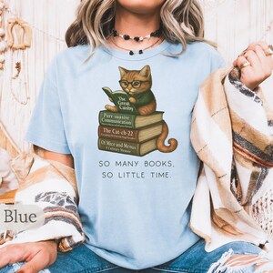 K&ouml;nnte beinhalten: Hellblaues T-Shirt mit einem Motiv eines Katers, der ein Buch mit dem Titel "The Great Gatsby" liest. Der Kater sitzt auf einem Stapel B&uuml;cher mit Titeln wie "Purr-suasive Communication", "The Cat-ch-22" und "Of Mice and Menus". Der Text "SO MANY BOOKS, SO LITTLE TIME." ist unter dem Motiv gedruckt.