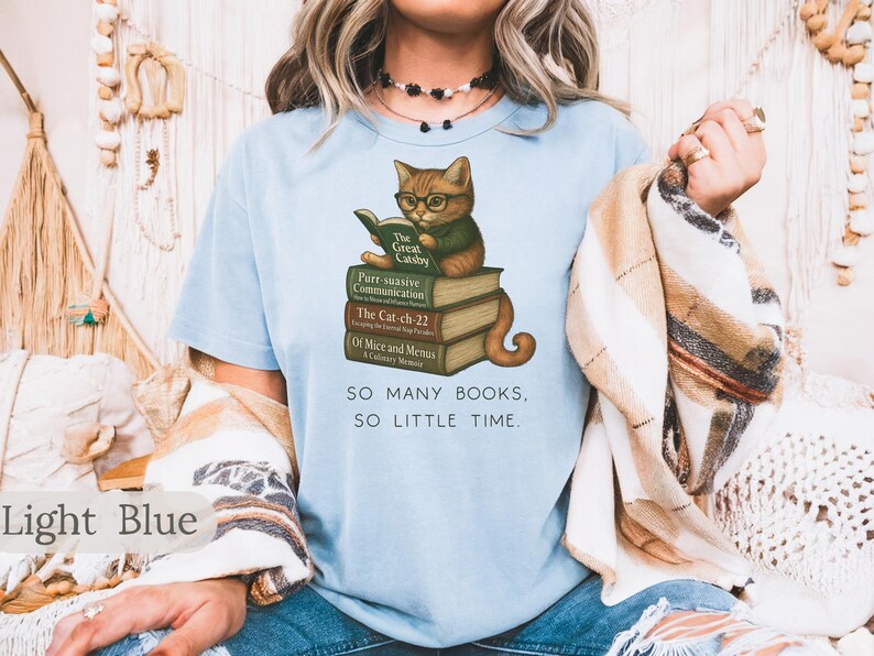 K&ouml;nnte beinhalten: Hellblaues T-Shirt mit einem Motiv eines Katers, der ein Buch mit dem Titel "The Great Gatsby" liest. Der Kater sitzt auf einem Stapel B&uuml;cher mit Titeln wie "Purr-suasive Communication", "The Cat-ch-22" und "Of Mice and Menus". Der Text "SO MANY BOOKS, SO LITTLE TIME." ist unter dem Motiv gedruckt.