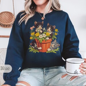 May include: A navy blue sweatshirt with a graphic of three kittens in a flower pot. The kittens are peeking out of the pot and surrounded by flowers.