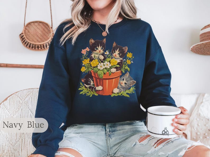 May include: A navy blue sweatshirt with a graphic of three kittens in a flower pot. The kittens are peeking out of the pot and surrounded by flowers.