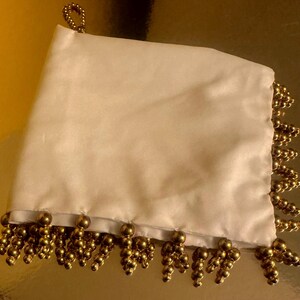 May include: Ivory satin pouch with gold beaded fringe. The pouch has a loop at the top for hanging. The gold fringe is made of small, round beads clustered together. The pouch is likely used for carrying small items.