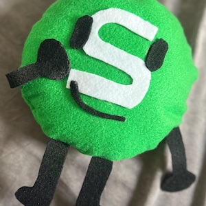 May include: A green, round plush character with a white 'S' on its front. It features black arms, legs, and a curved mouth. The soft fabric character has a playful, cartoonish design, suitable for children.