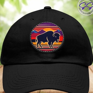 May include: Black baseball cap with a round patch featuring a silhouette of a buffalo standing in front of a sunset with mountains in the background. The patch has a stitched border and the text "Sips and Shenanigans" is visible in the upper right corner of the image.
