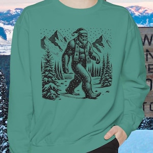 May include: Teal sweatshirt featuring a black and white illustration of Bigfoot wearing a Santa hat and scarf, walking through a snowy forest. The design includes mountains and pine trees, evoking a winter theme.