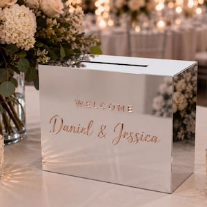 Op de afbeelding: Een rechthoekige spiegelende kaartenbox met een gleuf aan de bovenkant, voorzien van de roségouden tekst "WELCOME" en "Daniel & Jessica" in een sierlijk lettertype. Het reflecterende oppervlak toont de omgeving met bloemen en kaarsen.