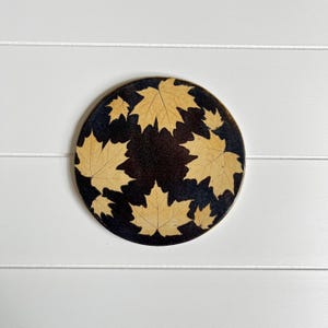 May include: A round coaster with a dark background and a pattern of light brown maple leaves. The coaster has a dark brown border and is set against a white, horizontal-slatted background.