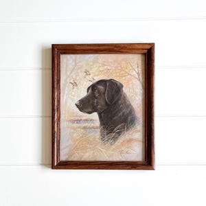 May include: A framed print featuring a black Labrador Retriever in a natural setting. The dog is depicted in profile, with a background of trees, water, and tall grass. The artwork is encased in a dark brown wooden frame, suitable for home decor.