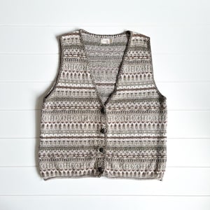 May include: A knitted vest with a V-neck and button closure. The vest features a geometric pattern in shades of brown, green, and cream. The garment is displayed on a white wooden surface.