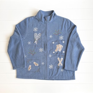 May include: A light blue fleece zip-up jacket with embroidered winter sports-themed patches. The patches feature snowshoes, ice skates, mittens, and skis, along with snowflakes and berry sprigs. The jacket has a high collar.