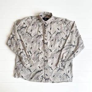 May include: A long-sleeved button-down shirt with a light tan background and a repeating pattern of fish, fishing nets, and other fishing-related graphics in black. The shirt has a button-down collar and a single chest pocket.