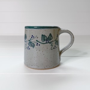 May include: A speckled ceramic mug with a light gray base and a dark green rim. The exterior features a hand-painted vine pattern with dark green leaves and small dark blue berries encircling the upper portion of the cylindrical body.
