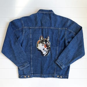 May include: A blue denim jacket with a detailed embroidered wolf head patch on the back. The wolf patch features shades of white, black, orange, and brown. The jacket has a classic button-up design with pockets and a collar.