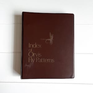 May include: A brown leather-bound book titled "Index of Orvis Fly Patterns" with gold lettering and a fly illustration. The book is rectangular and appears to be vintage, with some wear visible on the cover.