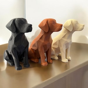 Può includere: Tre sculture geometriche di cani nei colori nero, marrone rossiccio e crema. Le statue dei cani sono sedute dritte con la testa girata di lato. Le sculture sono esposte su una superficie marrone chiaro.