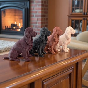 May include: Four decorative Cocker Spaniel figurines in a row, each in a different color: burgundy, black, terracotta, and cream. The figurines are sitting on a polished wooden surface, with a fireplace and cozy living room setting in the background.