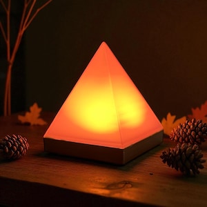 Op de afbeelding: Een piramidevormige lamp die een warm oranje en geel licht uitstraalt. De lamp staat op een houten oppervlak, met dennenappels en herfstbladeren. De lamp heeft een witte basis.