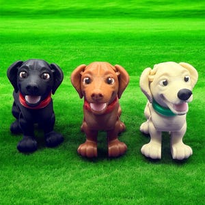 Puede incluir: Tres perros de juguete, uno negro, uno marrón y uno blanco, están de pie sobre una superficie verde cubierta de hierba. Todos miran a la cámara y tienen una expresión feliz en sus caras.