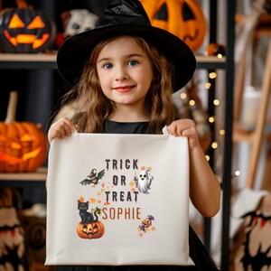 Puede incluir: Una niña con un sombrero de bruja negro sostiene una bolsa de Halloween blanca. La bolsa presenta un gato negro sobre una calabaza, un murciélago, un fantasma y las palabras "Trick or Treat Sophie". Hay calabazas naranjas en el fondo.